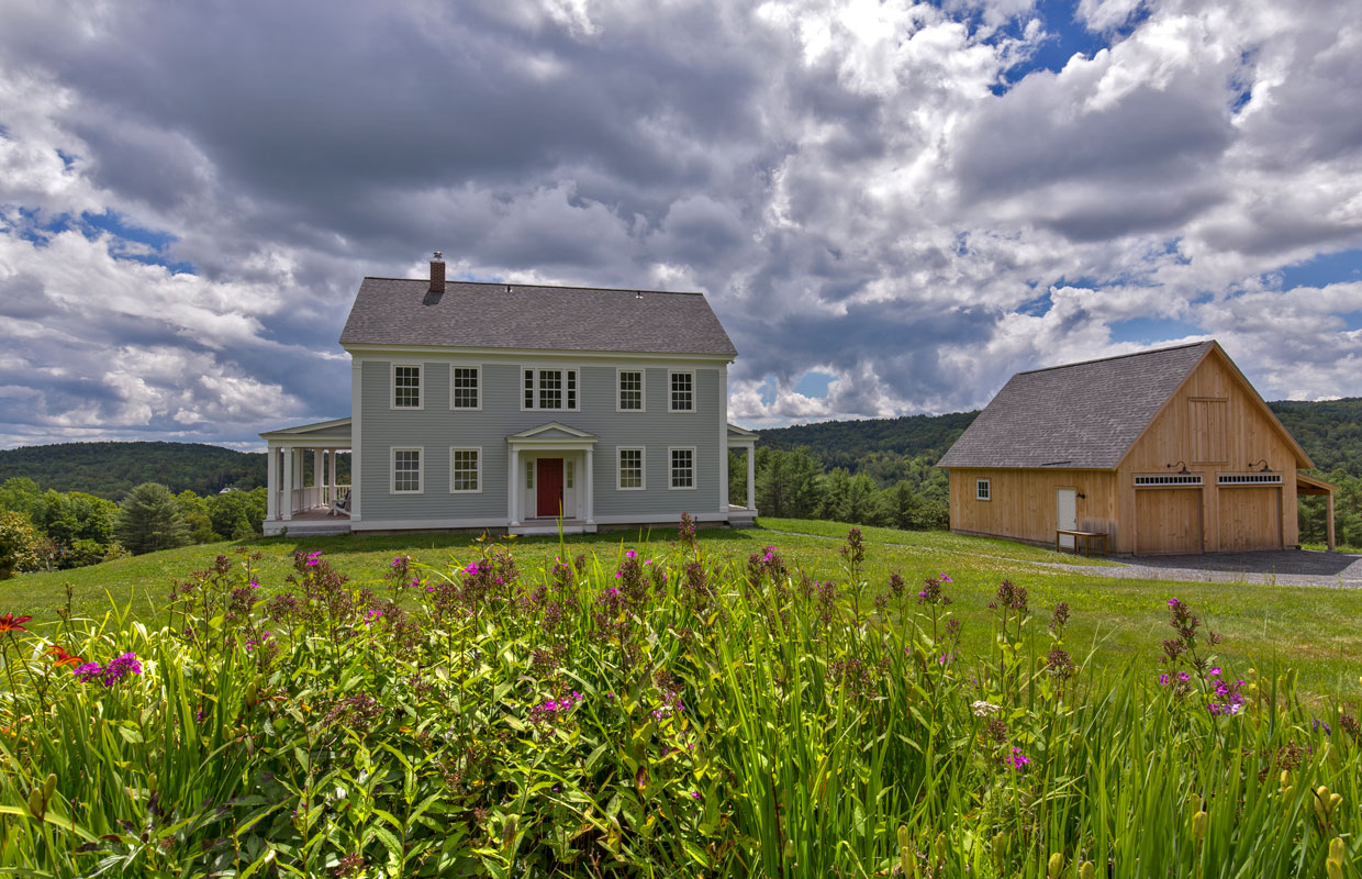 large gray colonial home on a hill with a barn to the right
