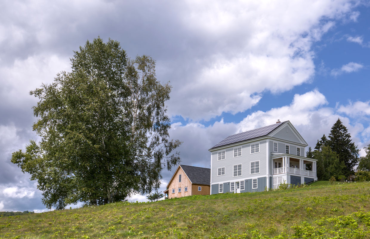 view from down a hill at a large gray colonial home on top of the hill with a barn to the left